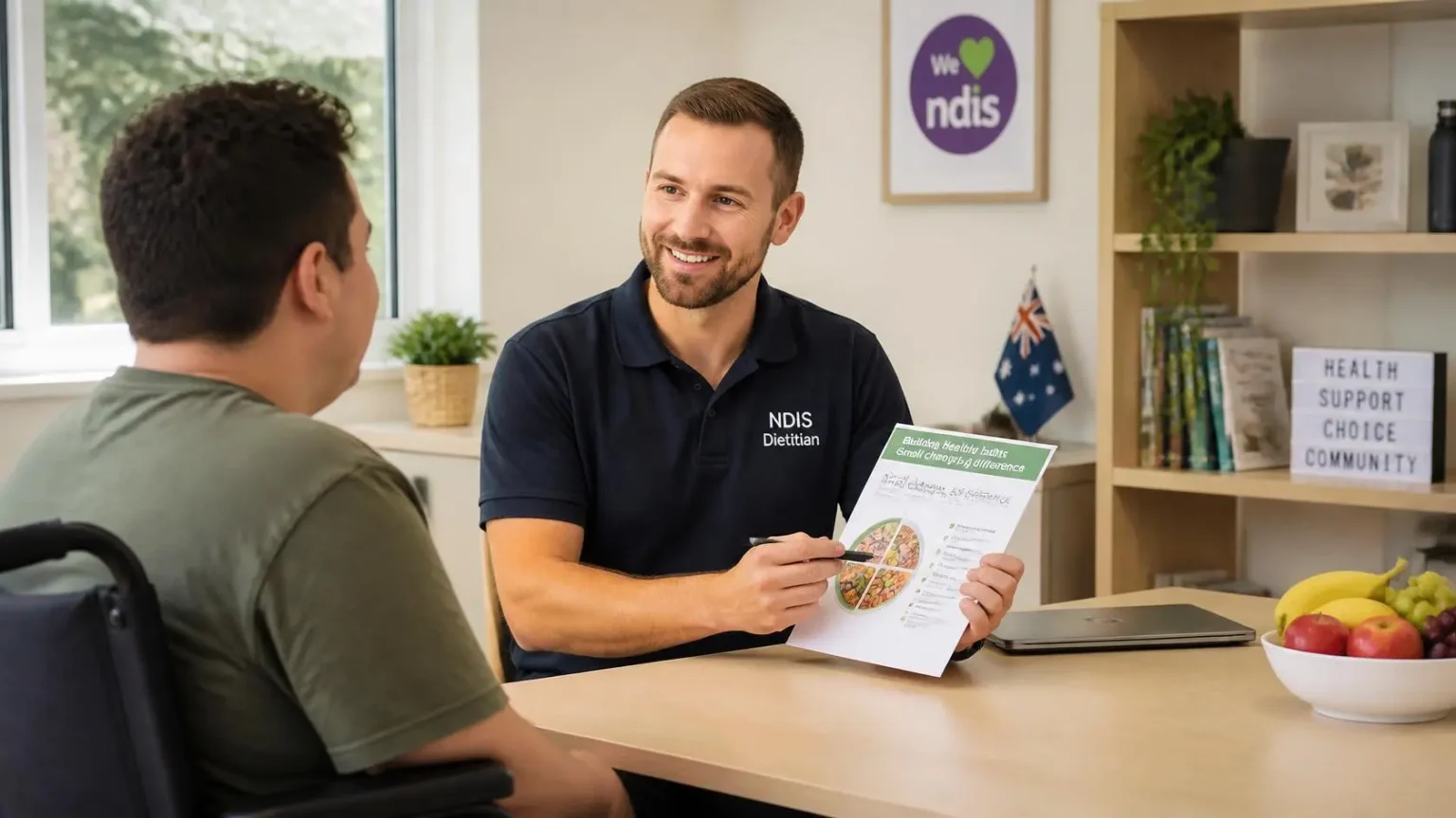 NDIS dietitian in a branded shirt taking consultation notes during an in-home meal planning assessment with a wheelchair participant.